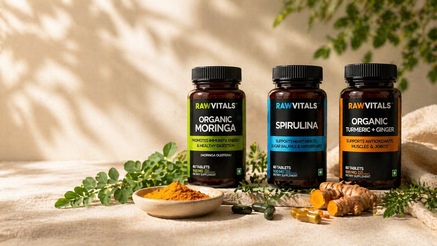 Three Rawvitals organic supplement bottles — Moringa, Spirulina, and Turmeric + Ginger — on a sunlit cream surface with fresh moringa leaves, turmeric powder and root, and capsules