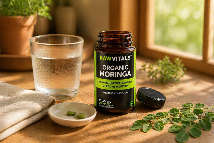 Rawvitals Organic Moringa on a sunlit kitchen counter with a glass of water, two tablets, fresh moringa leaves and a linen napkin